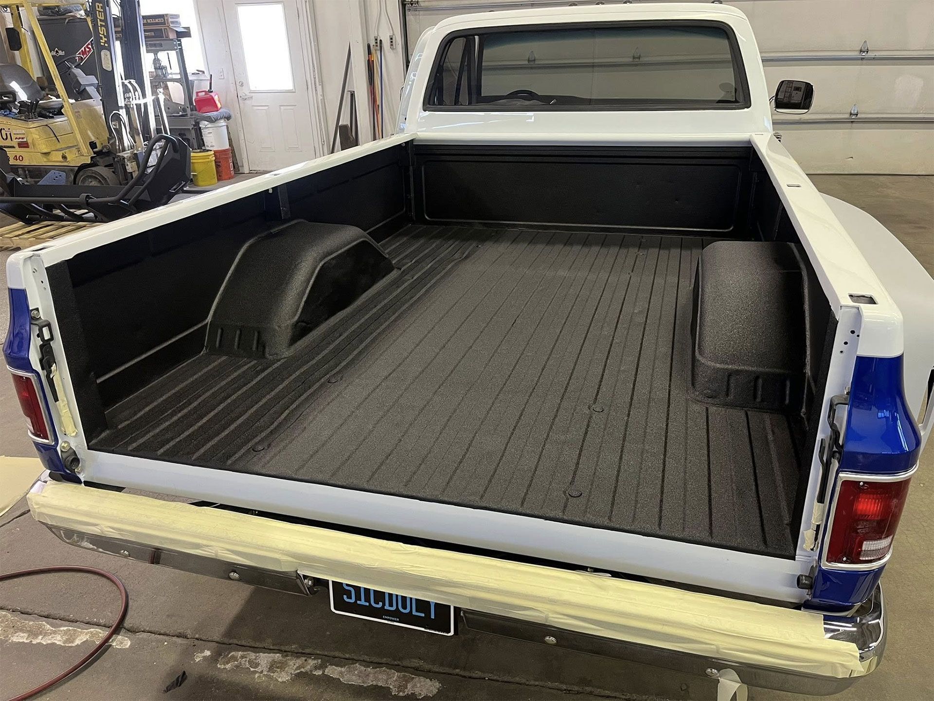 The rear view of a white pickup truck with a newly applied black bed liner, parked inside a workshop.