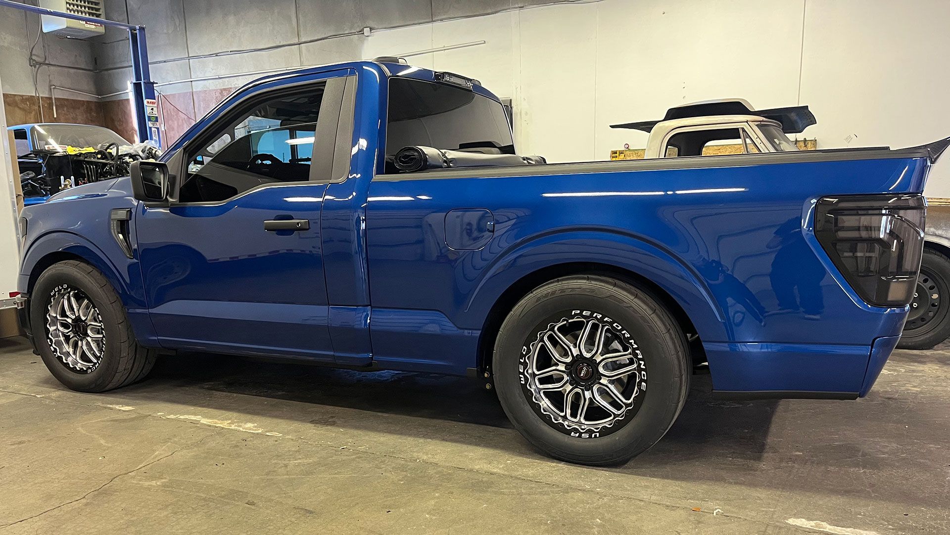 A blue regular cab pickup truck with aftermarket custom wheels parked inside a garage.