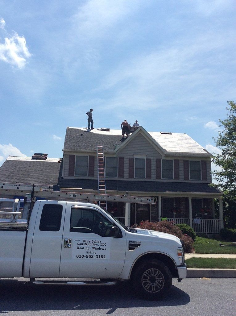 Workers on a house roof with a white truck in front; blue sky.