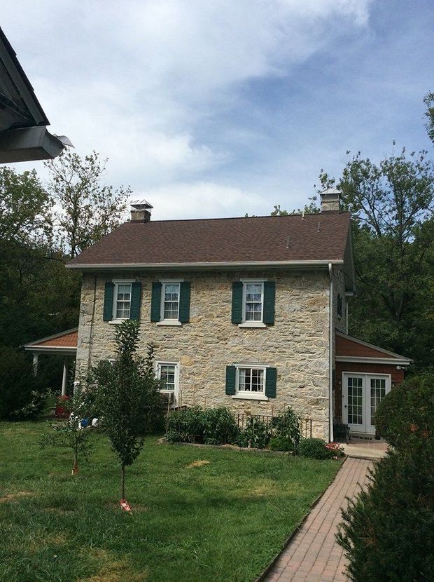Stone house with green shutters, brown roof, and brick walkway surrounded by greenery.