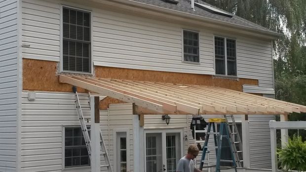 Construction of a wooden pergola attached to a two-story house with a person working on it.