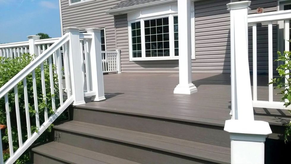 Wooden deck with stairs, white railings, and house with a bay window.