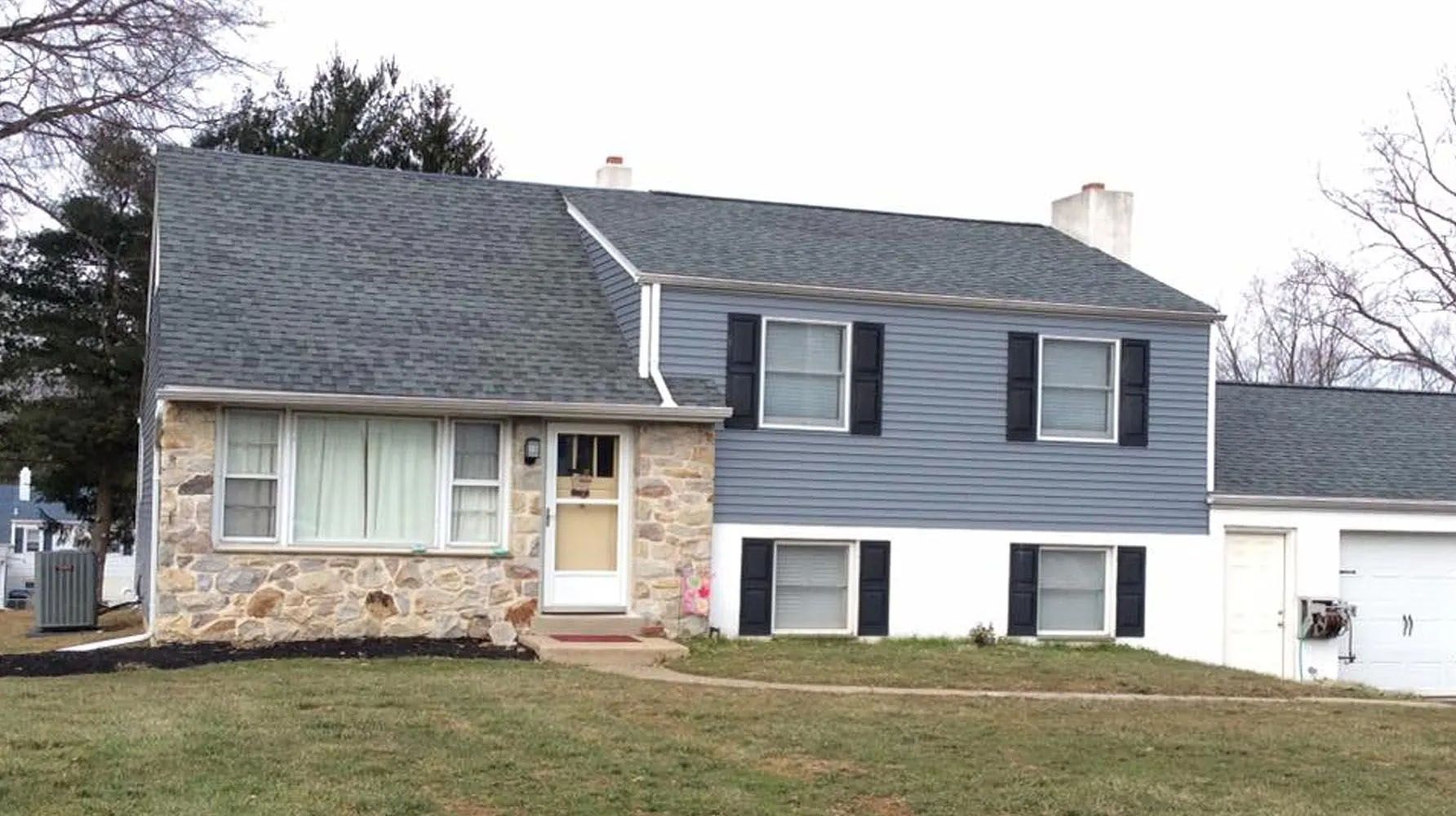 House with stone and blue siding, dark roof, black shutters, white garage door, and overcast sky.