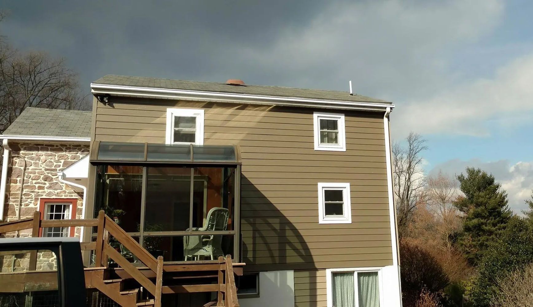 Two-story house with brown siding, screened porch, and wooden deck. Cloudy sky in background.