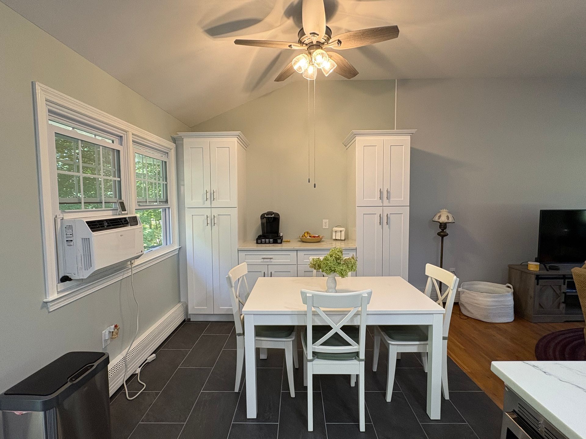 White kitchen with a table, chairs, and cabinets.  Gray tile floor, air conditioner, and a window.