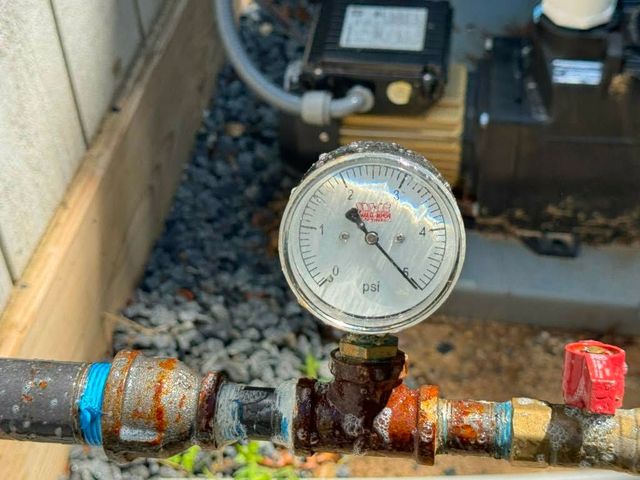 Pressure gauge on a rusty outdoor pipe with a red valve in a gravel-filled utility area