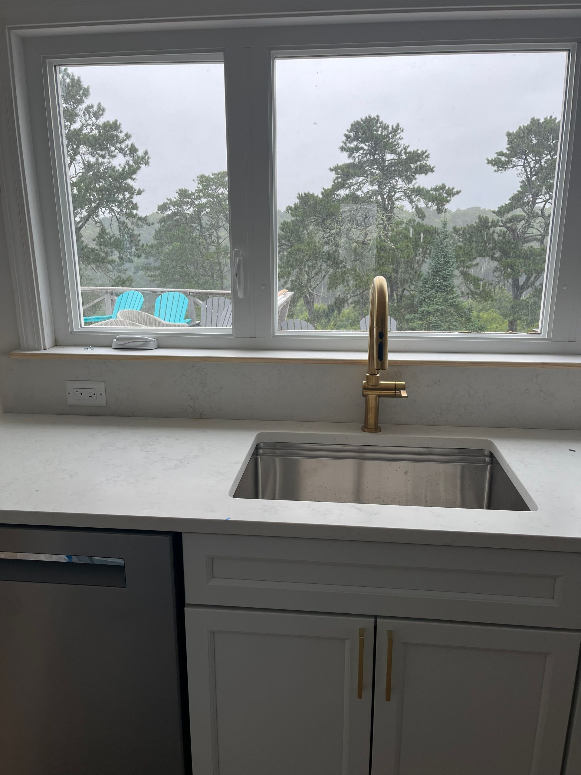 Kitchen sink under a large window with a gold faucet and white cabinets, overlooking trees outside