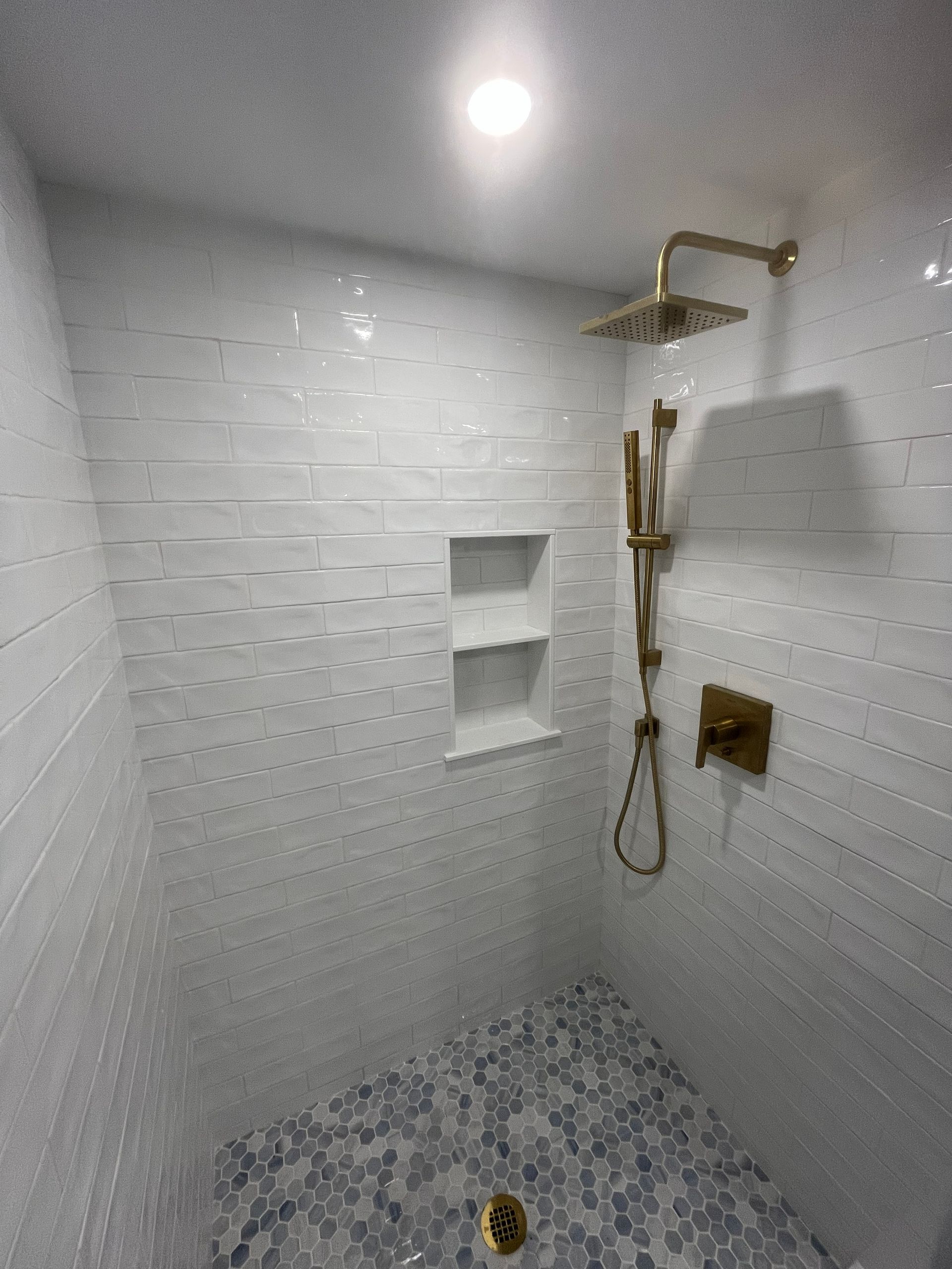 Modern white-tiled shower with brass fixtures, built-in shelf, and gray mosaic floor drain.