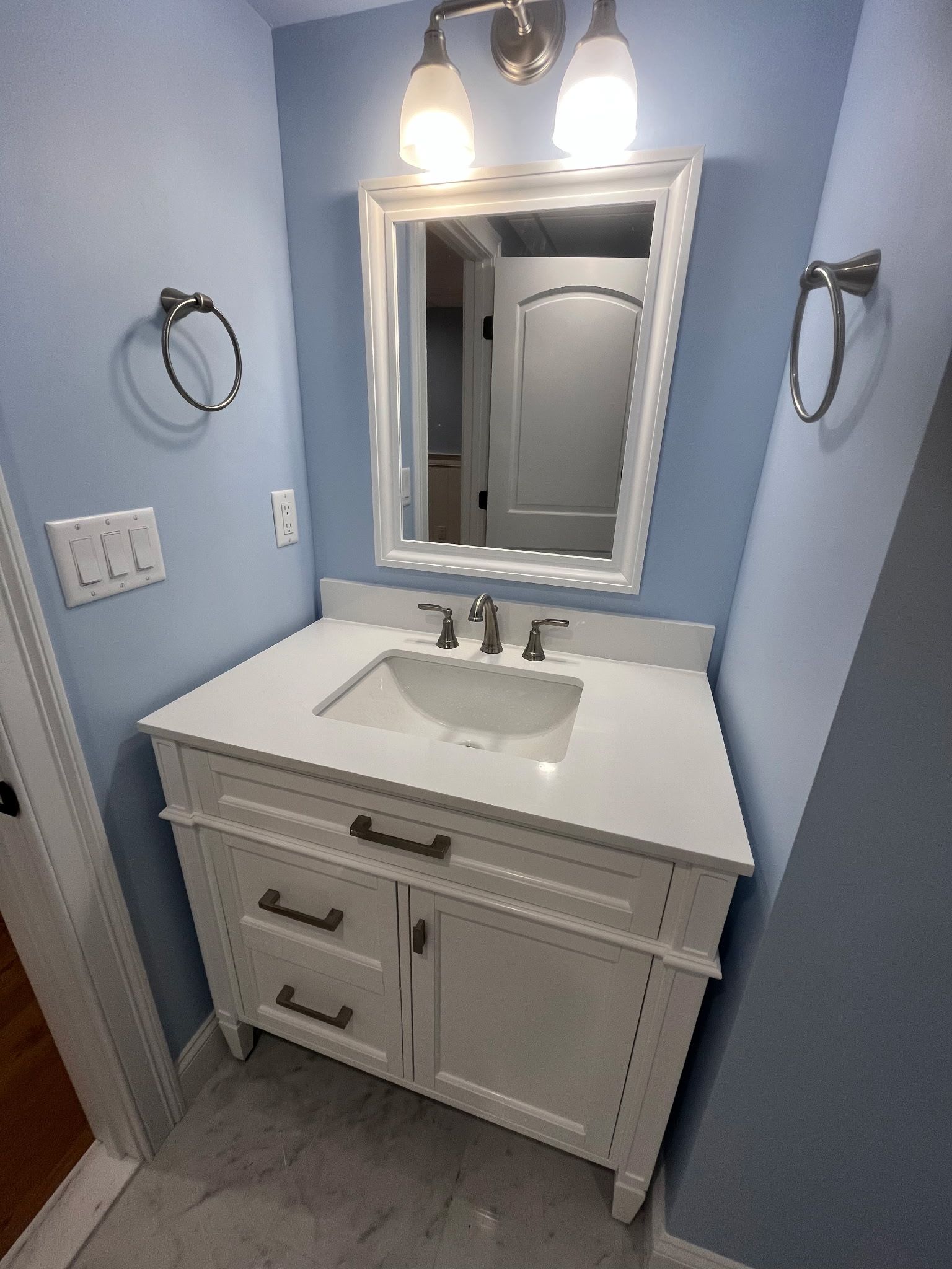 Small bathroom vanity with white sink, mirror, and light blue walls under a pair of lights.