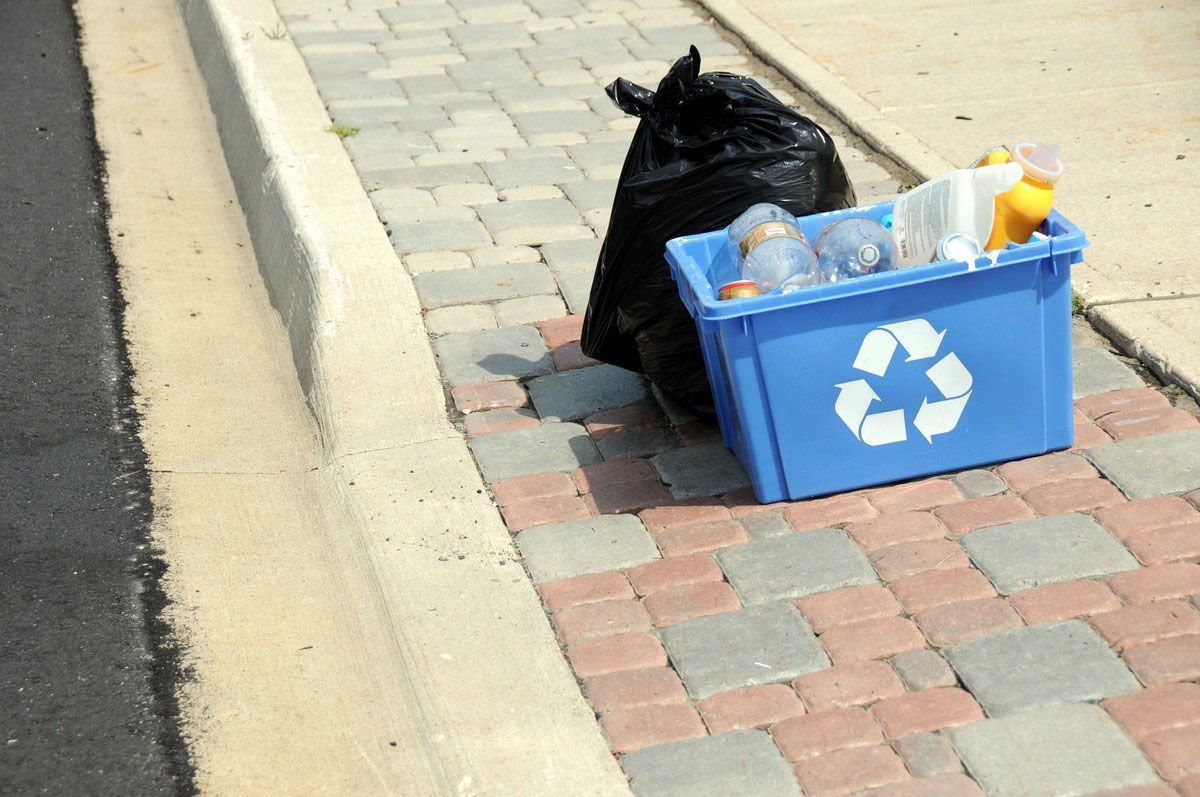 A black trash bag and blue recycling bin on a brick sidewalk next to a road.
