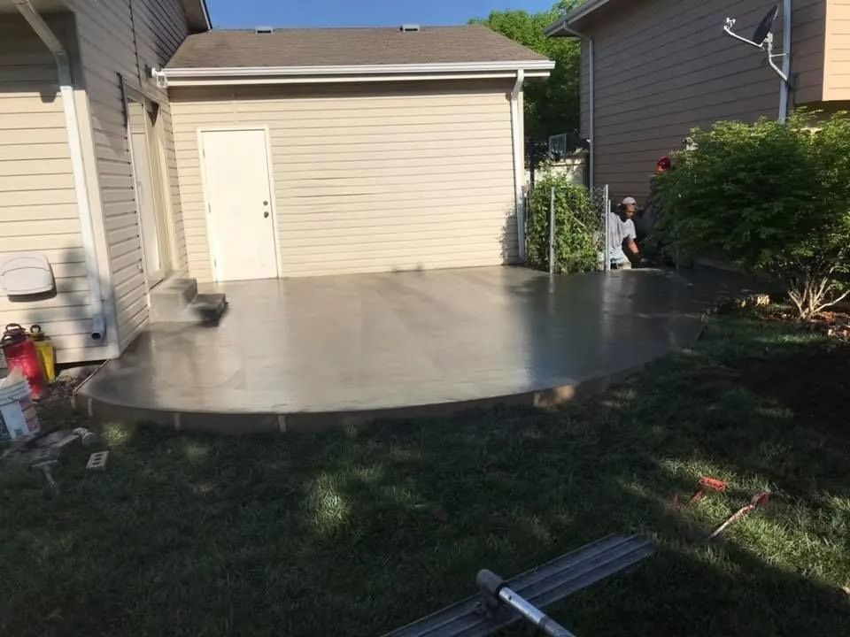 Newly poured concrete patio next to a light tan garage. A person kneels in the background. Green grass surrounds the patio.
