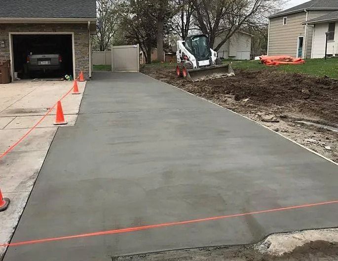 Newly poured concrete driveway with a garage on the left, an excavator in the background, and orange cones marking the perimeter.