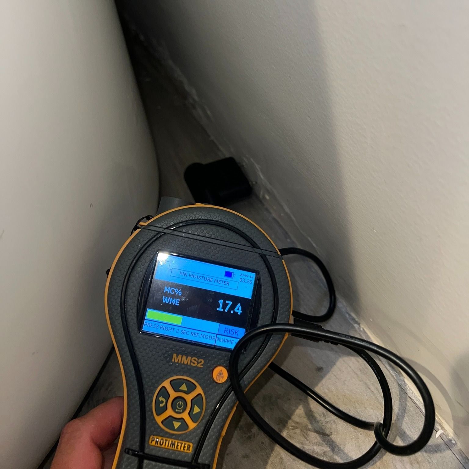 Moisture meter in a bathroom corner, reading 17.4. White wall and a black cable are visible.