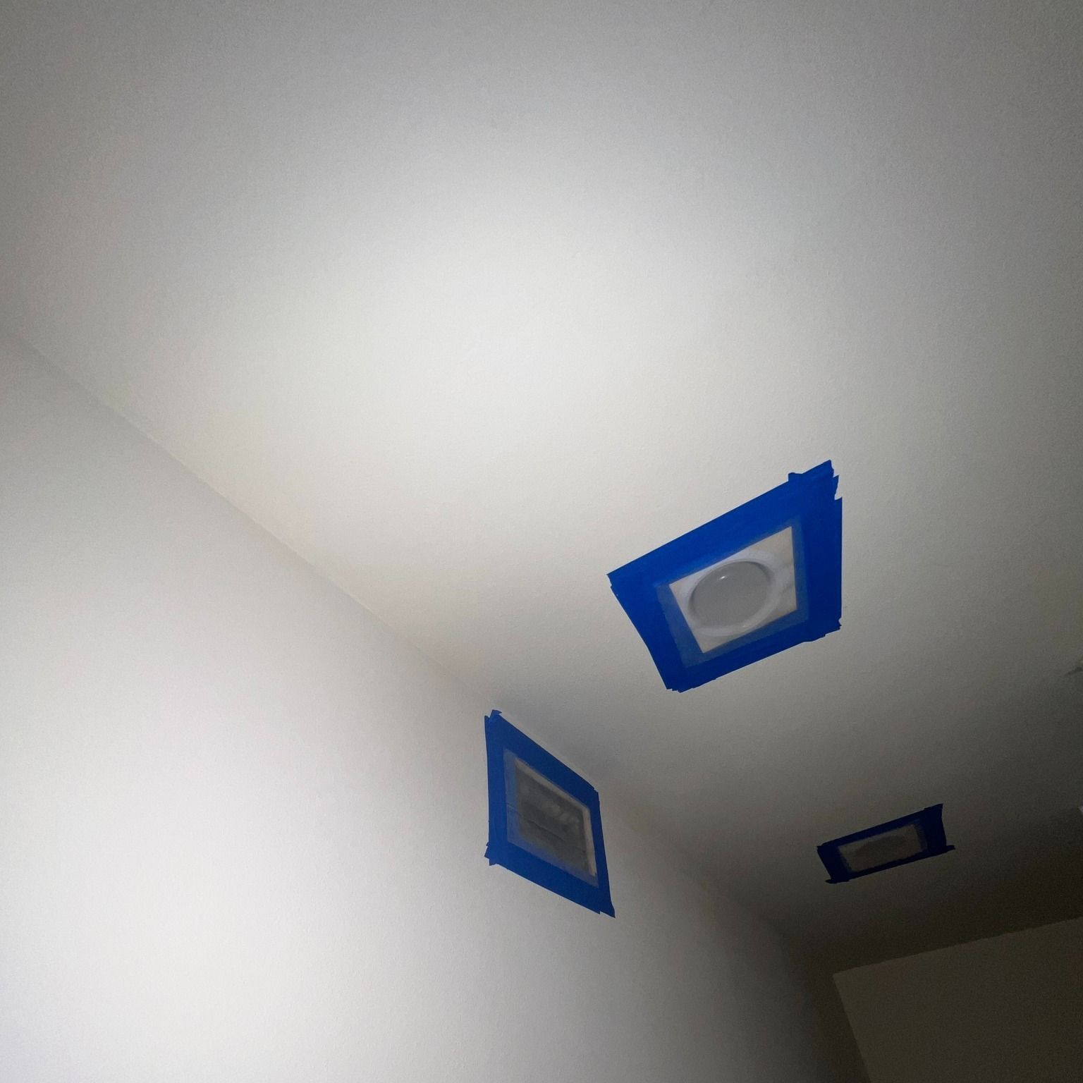 Three recessed ceiling lights taped off with blue painter's tape, in a white room.