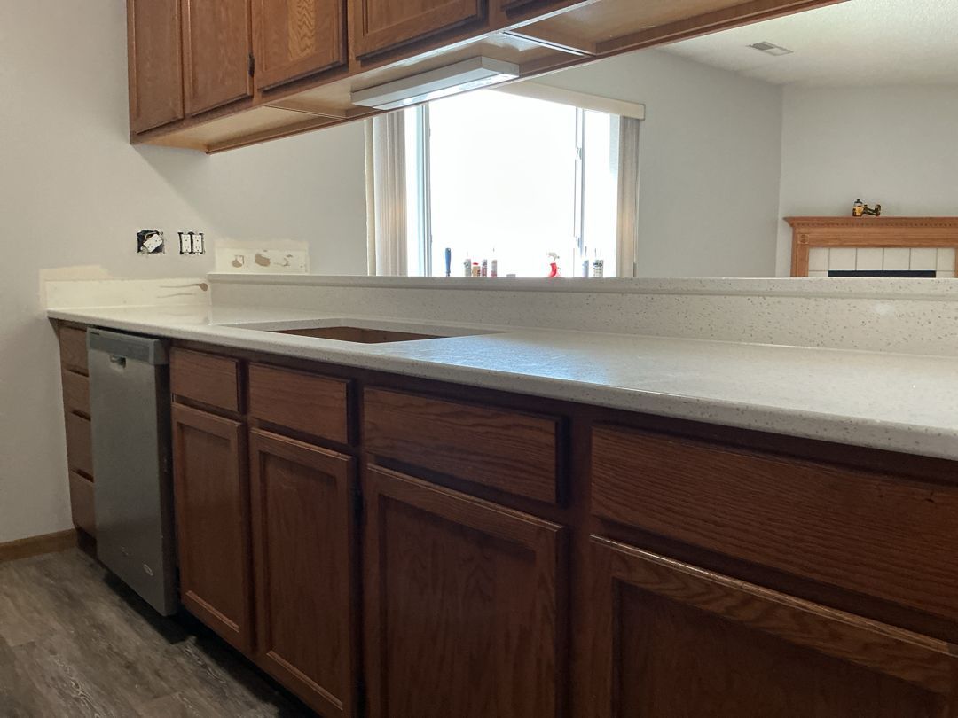 Kitchen with brown cabinets, white countertop, dishwasher, and window overlooking a living space.