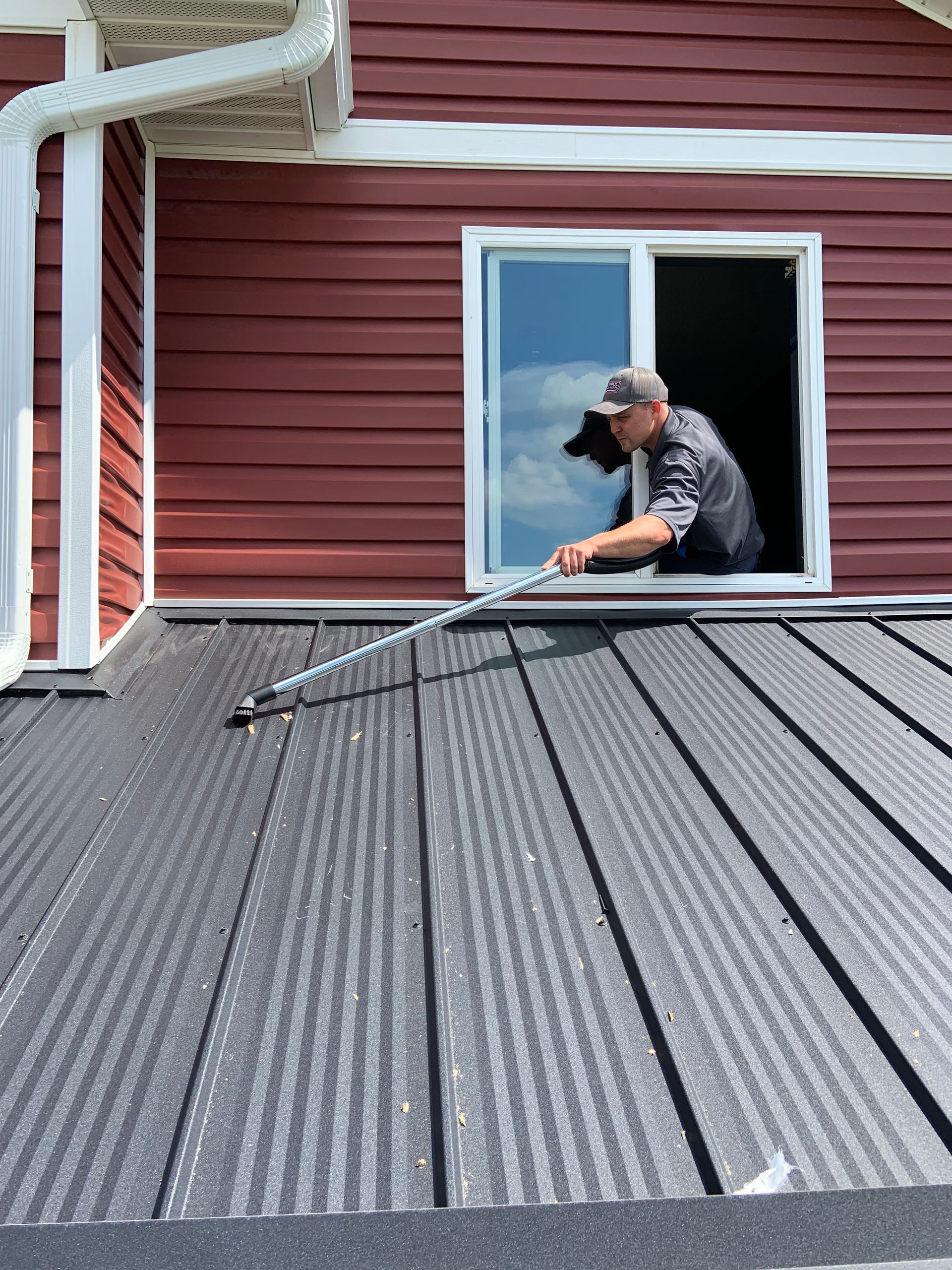 Person cleaning a dark metal roof from an open window of a red house.