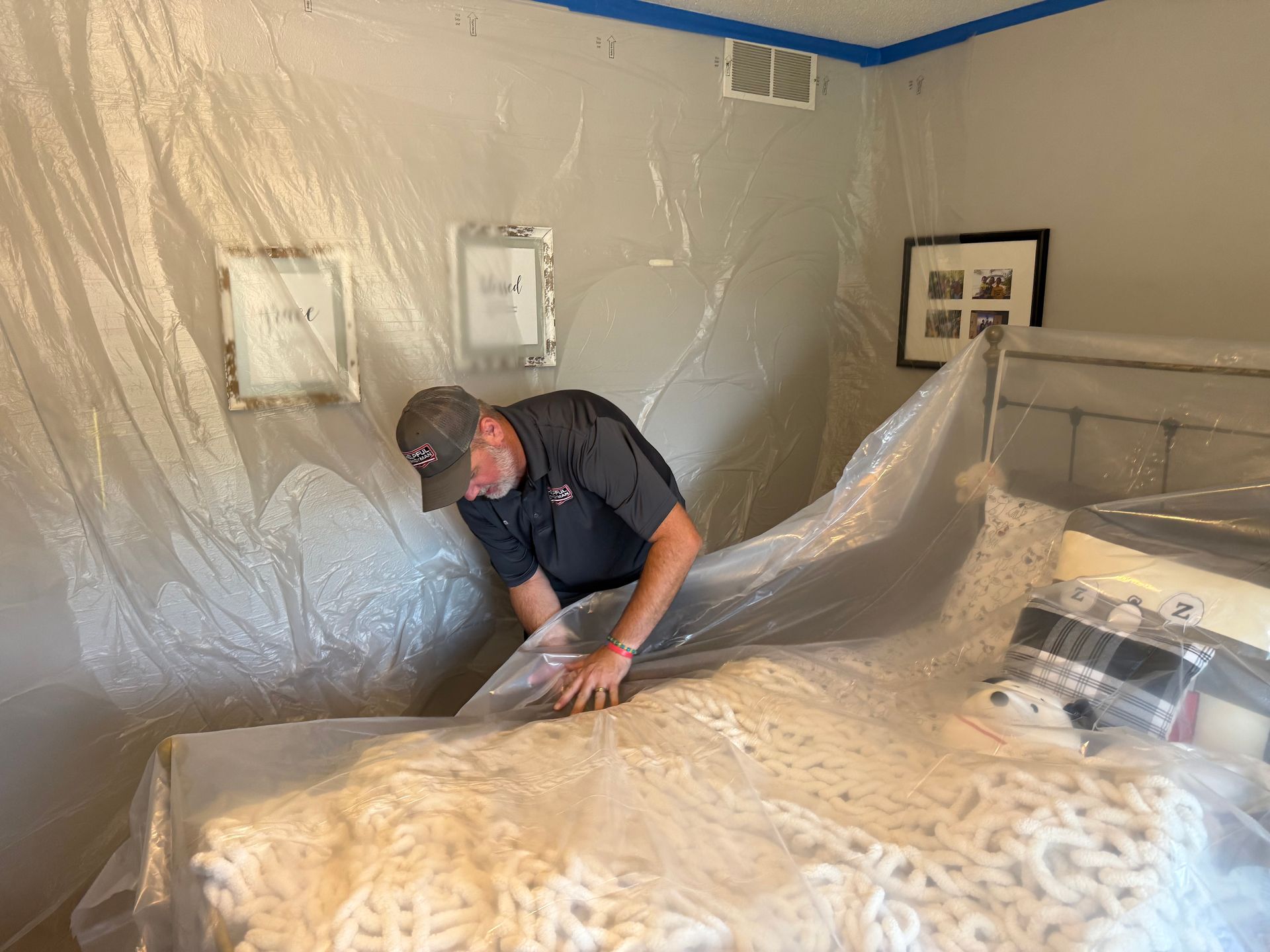 Man covering bed with plastic sheeting in a room, preparing for a project. Walls and pictures protected.