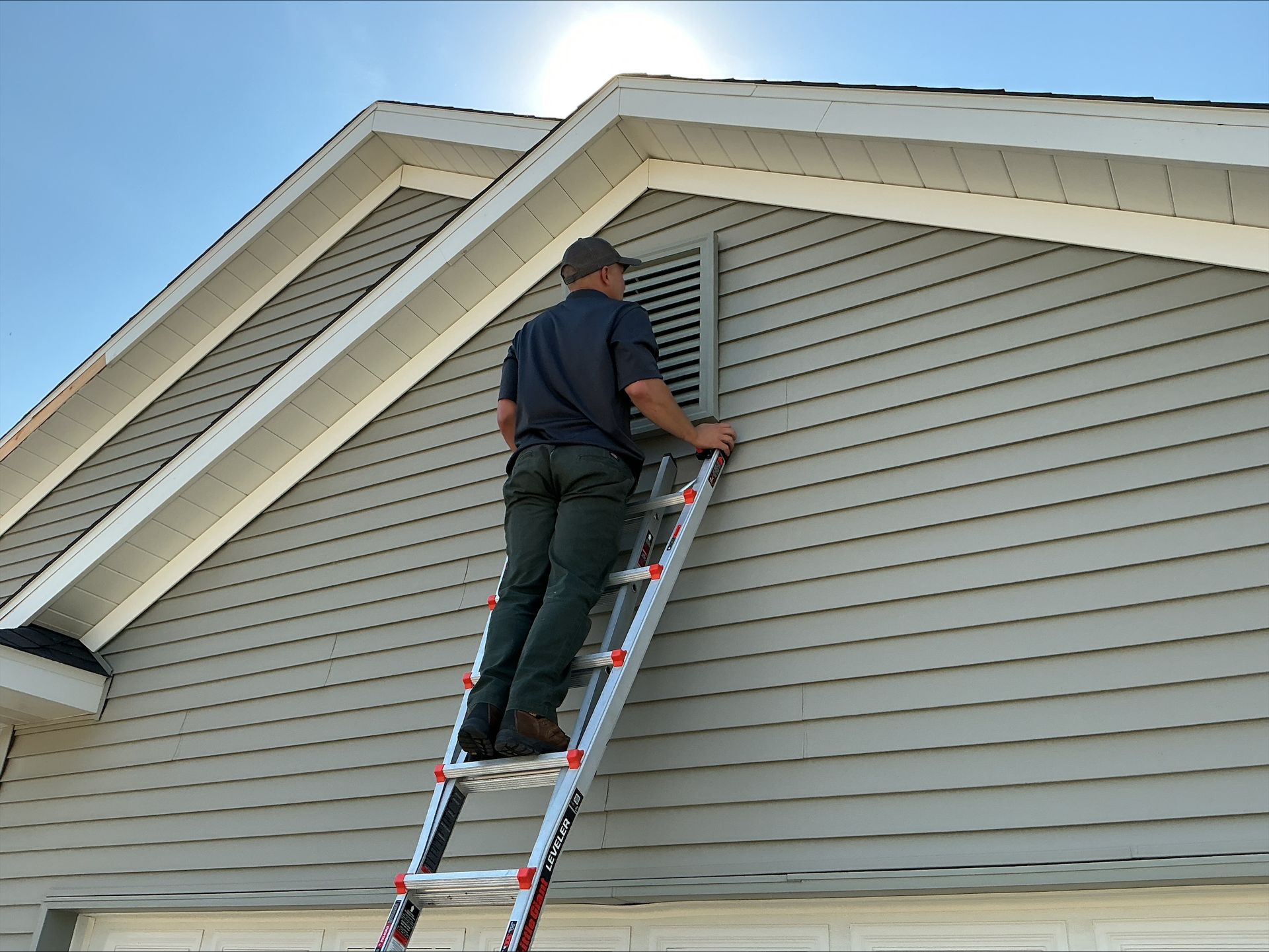 Person on ladder inspecting attic vent on a house with gray siding.
