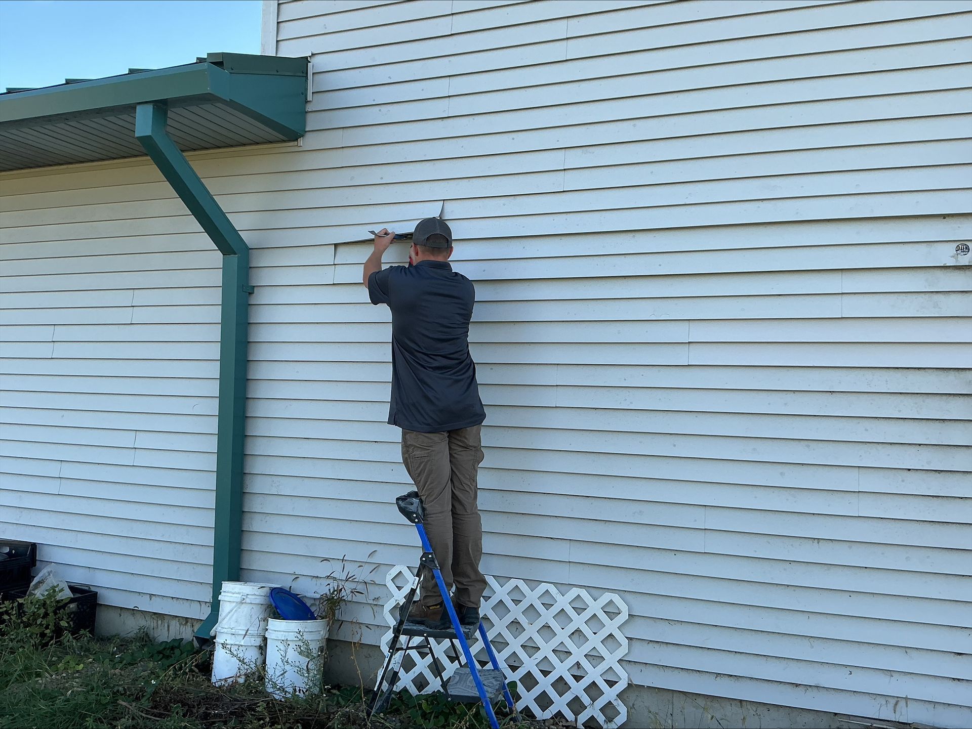 A person on a ladder is installing something on a white siding wall next to a green gutter.