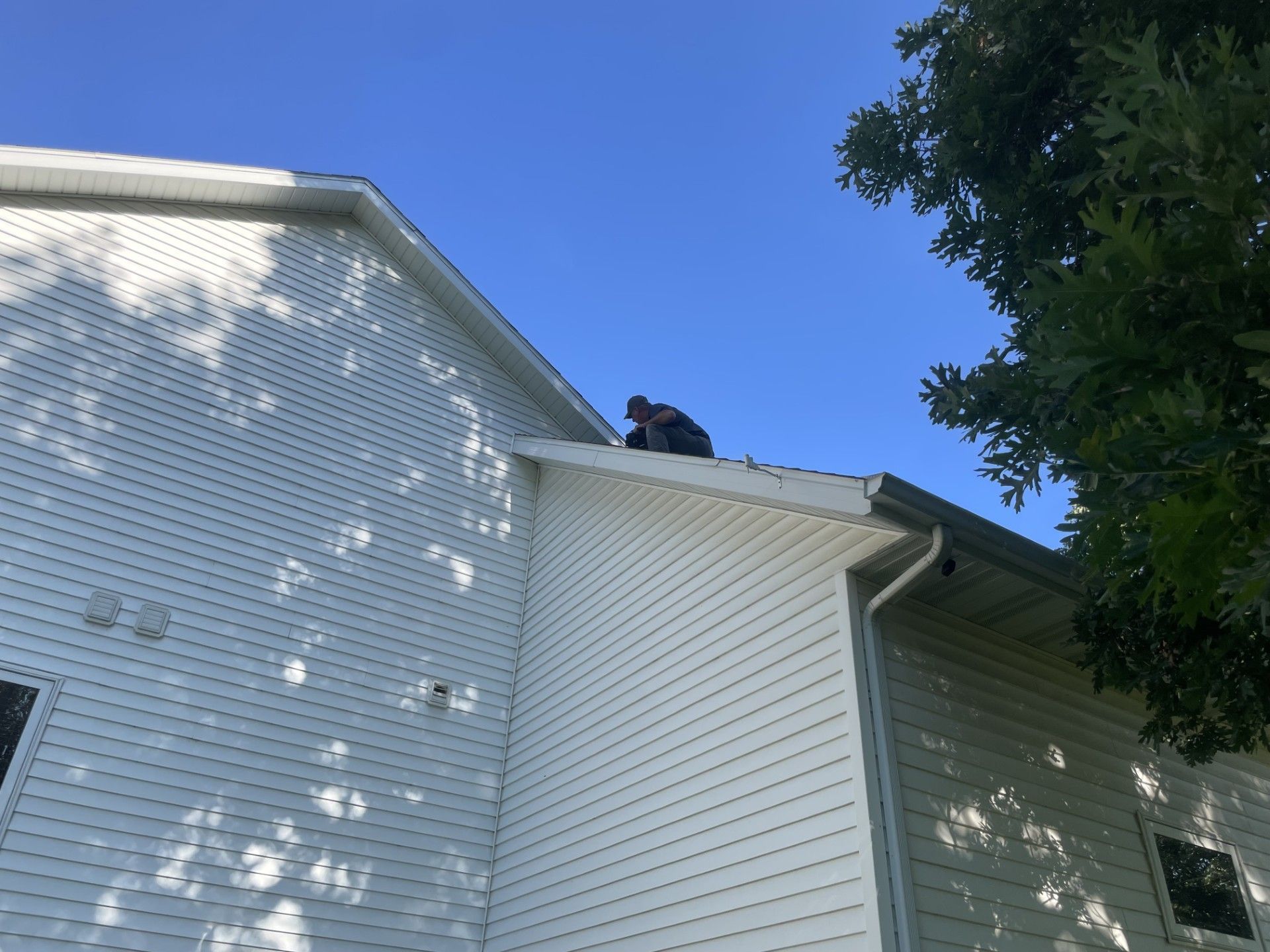 Person on a roof, inspecting or working. White siding, blue sky, tree branches.