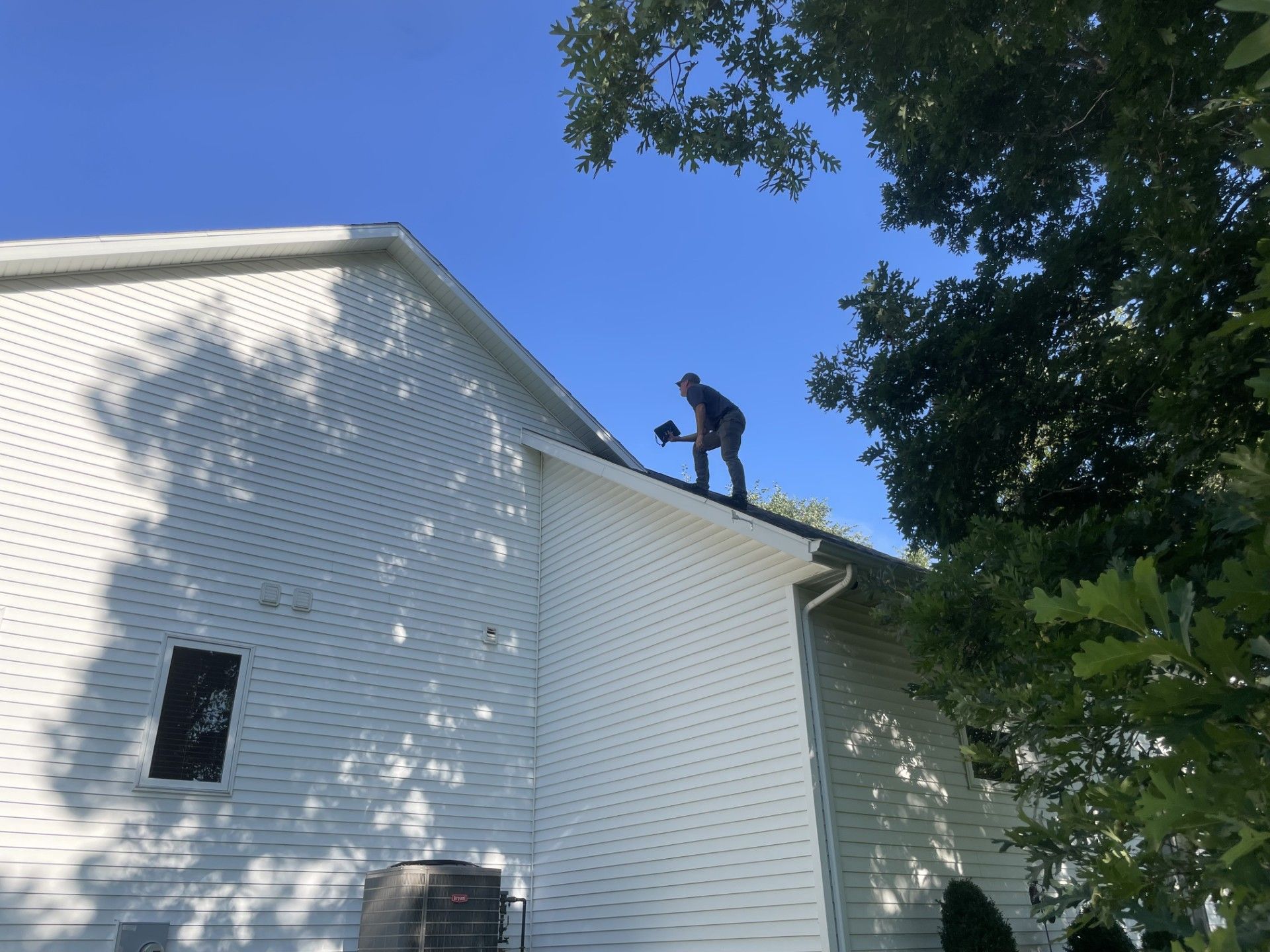 A person stands on a roof, cleaning gutters on a sunny day. White house, blue sky, green tree.
