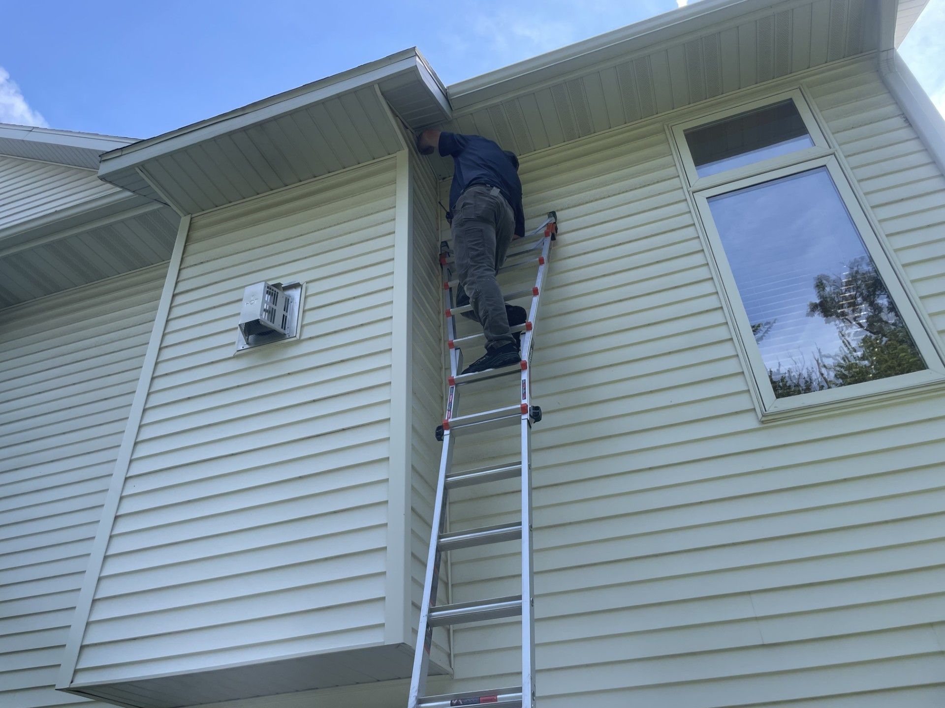 Person on ladder working on the side of a beige house with a window.