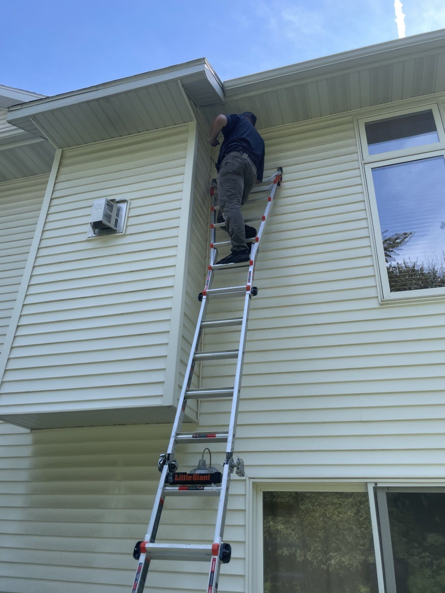 Man on a ladder, working on a house's gutter. White siding, clear sky.
