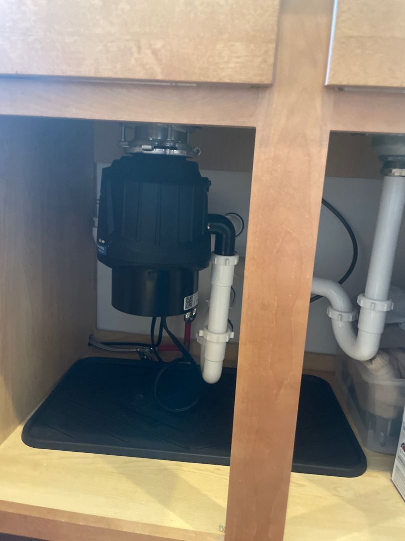 Black garbage disposal unit under a kitchen sink. White plumbing and black mat are visible.