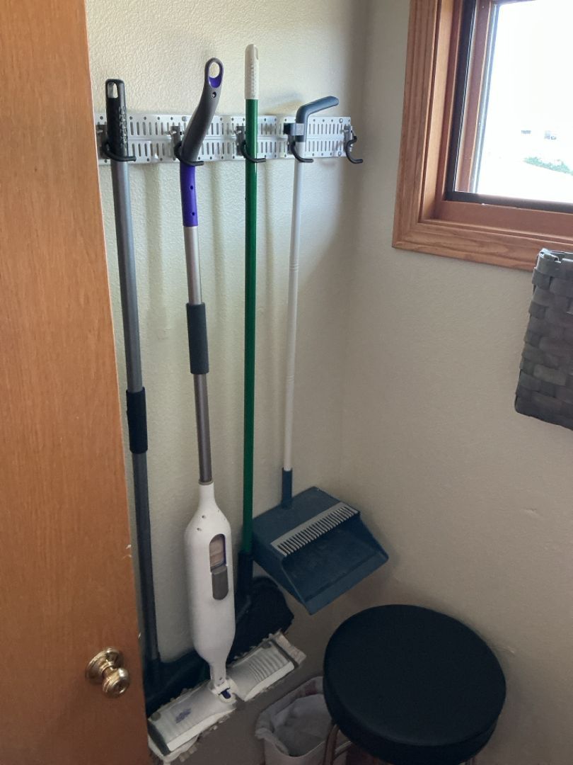 Cleaning tools hanging on a wall-mounted rack next to a door and window. Includes mops, brooms, and dustpan.