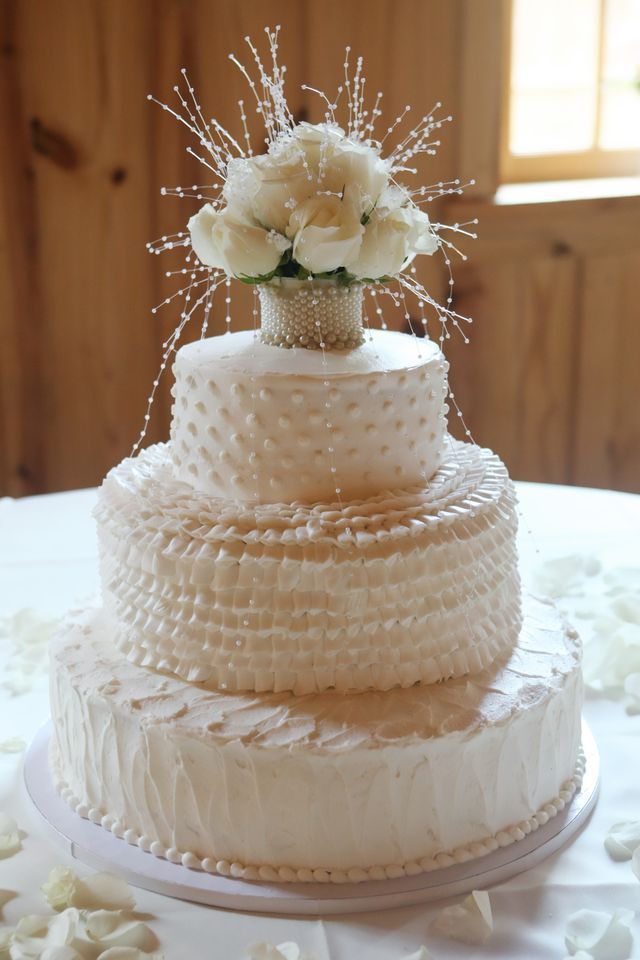 A three-tiered white wedding cake decorated with ruffled texture, pearls, and a floral topper with decorative wire accents.