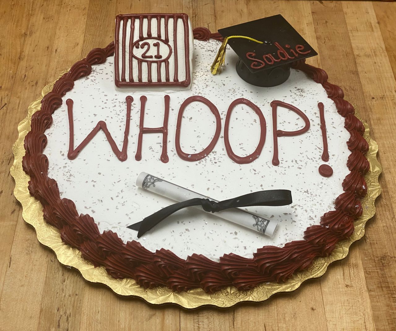 Round celebration cake with red icing border, WHOOP! written on top, and a small graduation cap decoration.