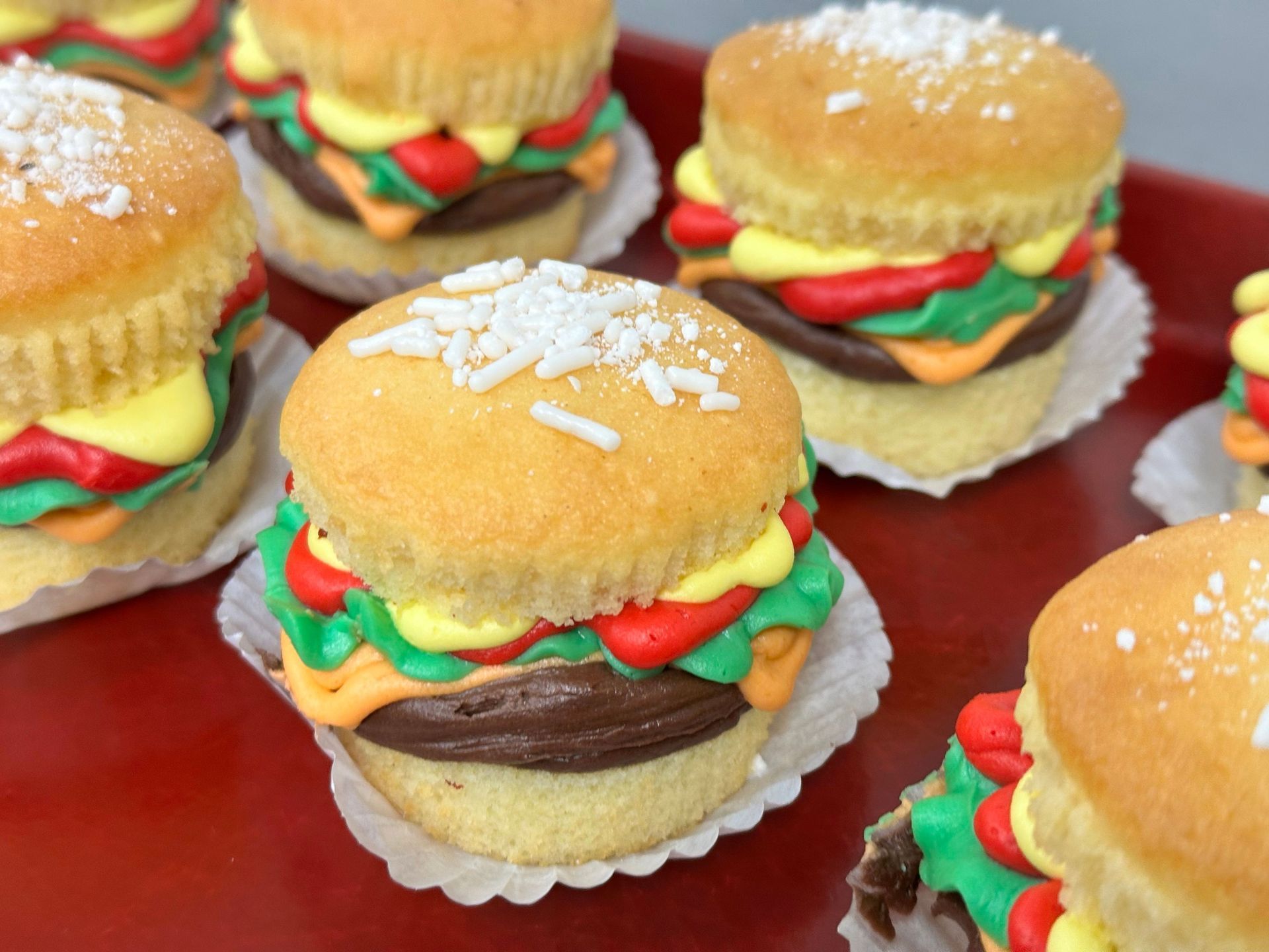 Mini cupcakes decorated like cheeseburgers on a tray, with sesame seeds and colorful frosting toppings