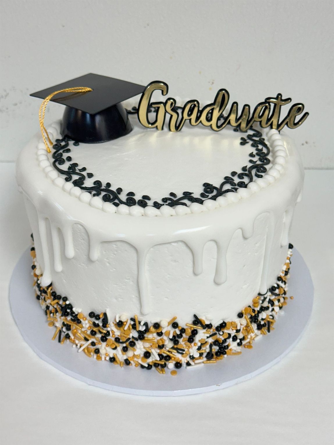 White graduation cake with black-and-gold drip, cap topper, and Graduate decoration