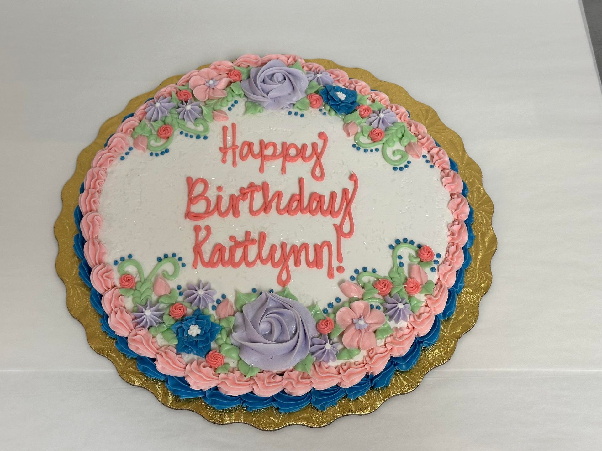 Birthday cake with pink floral icing and gold scalloped edge, reading “Happy Birthday Kaitlynn!”