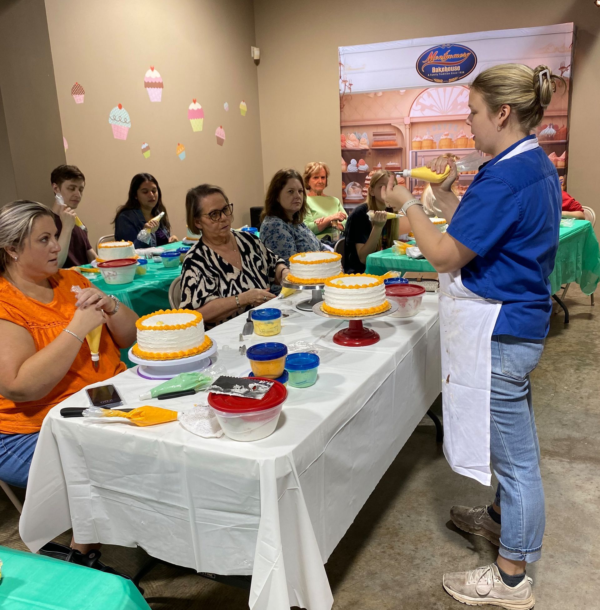 Cake decorating class: Instructor demonstrates piping. Students watch, working on cakes at a long table.