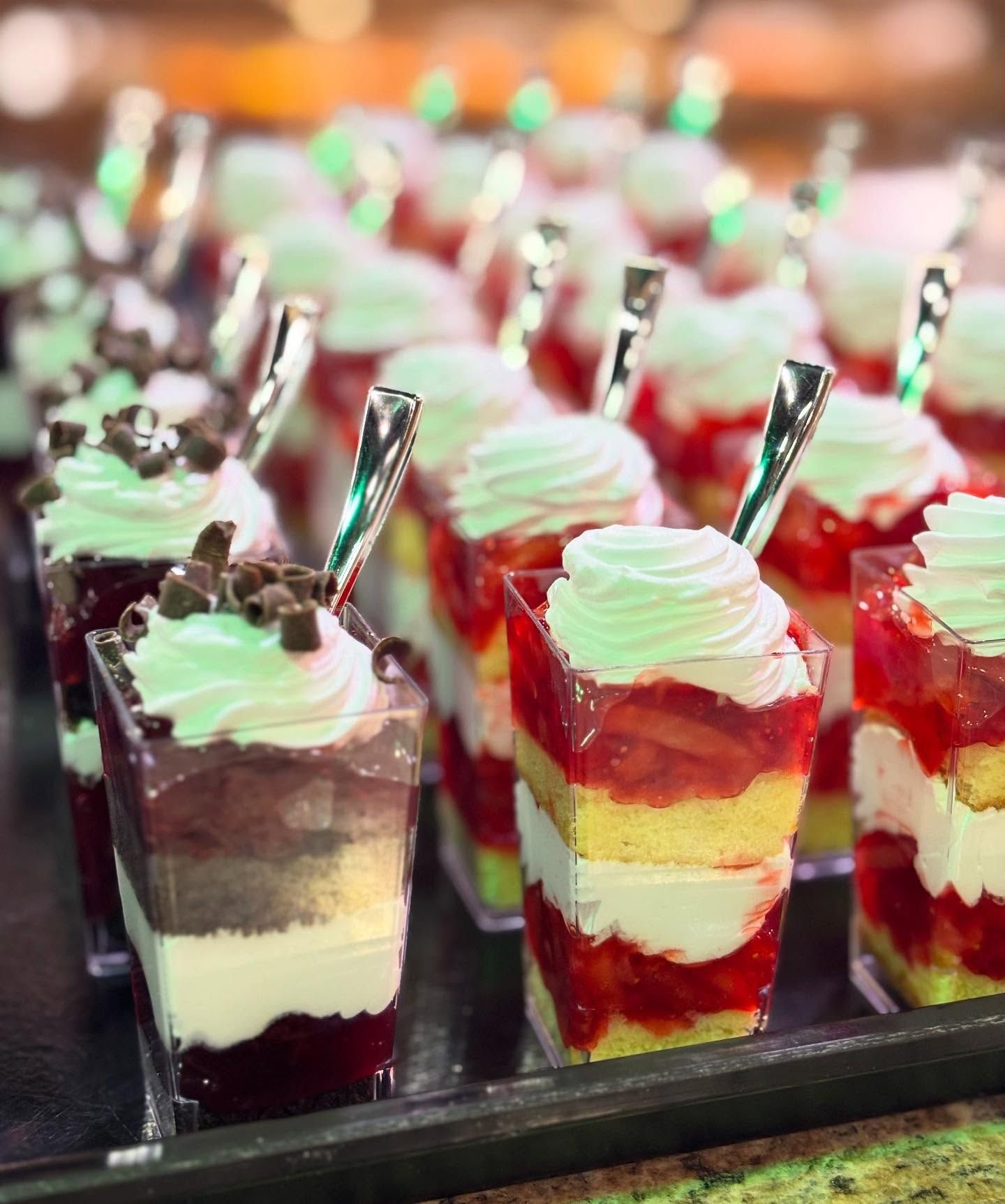 Rows of colorful layered desserts in clear cups, topped with whipped cream and a tiny spoon.