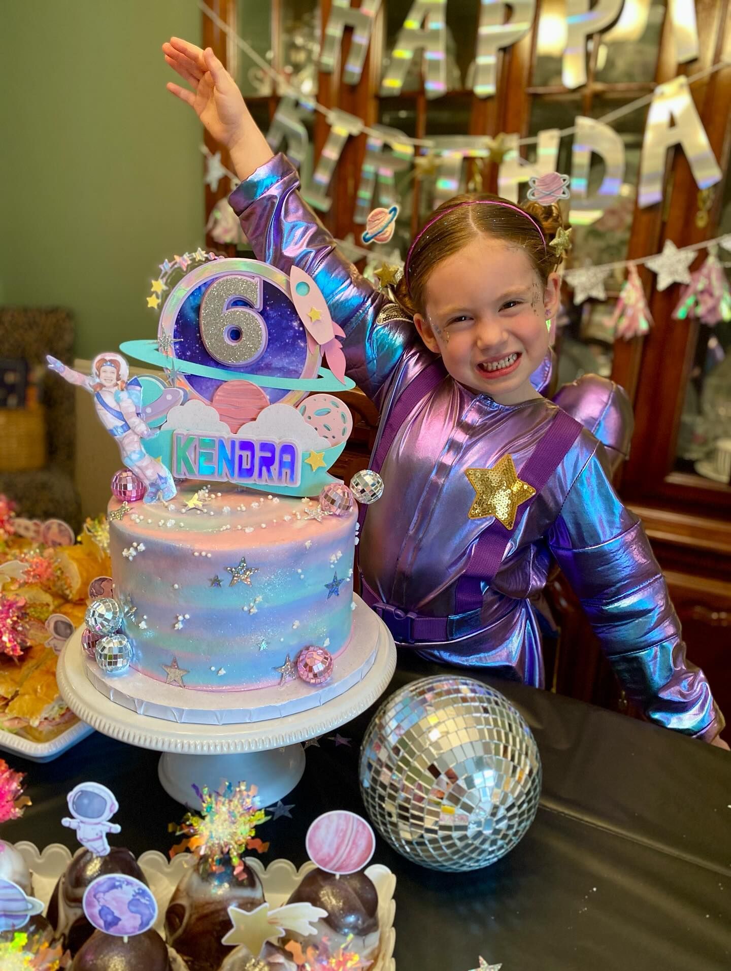 Girl in space suit celebrates her 6th birthday with a galaxy-themed cake, arm raised, smiling.