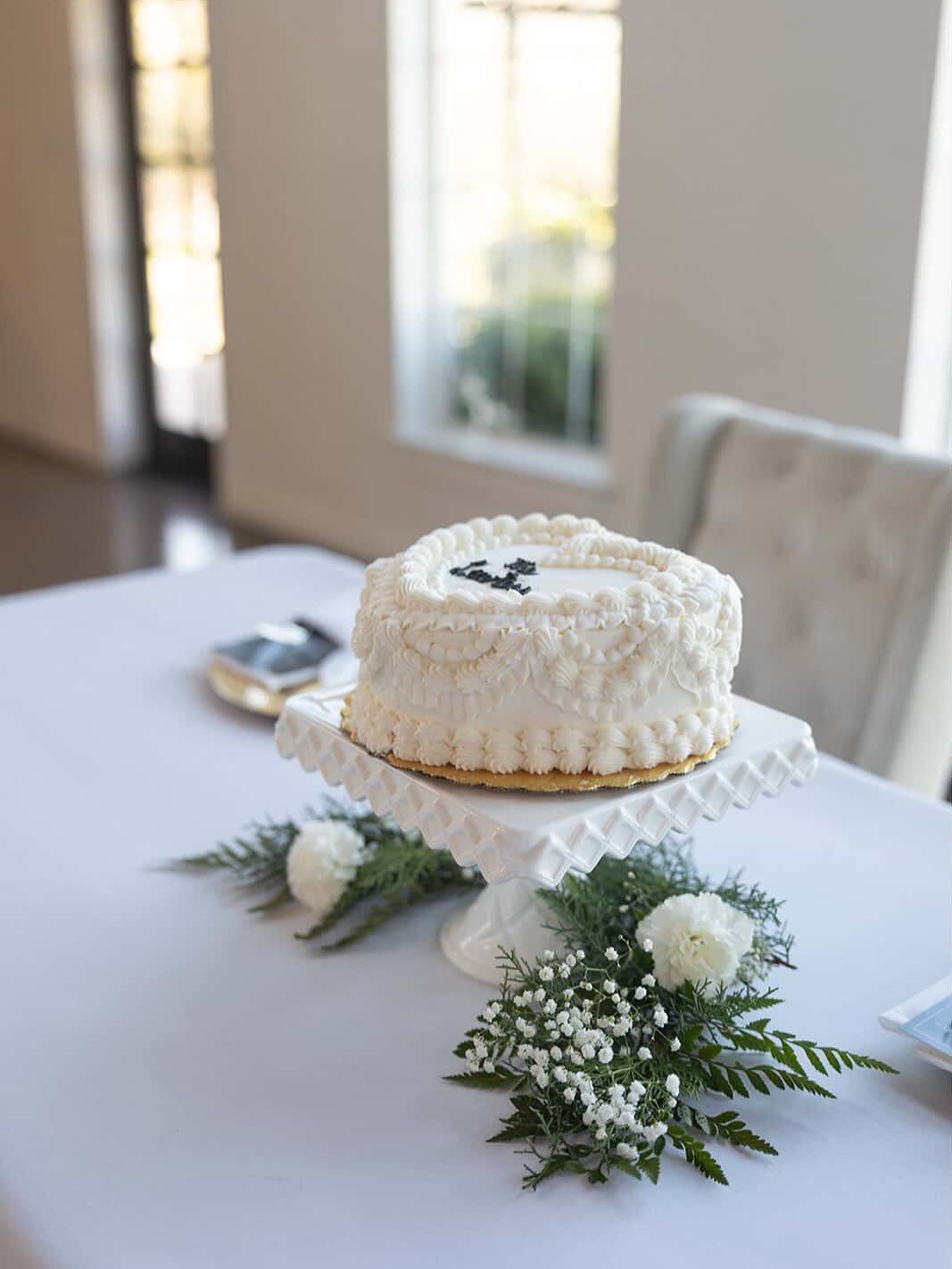 White frosted cake on a white pedestal, decorated with greenery, on a table in a well-lit room.