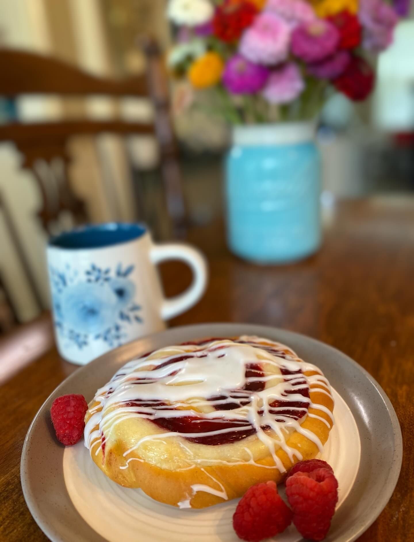 Raspberry pastry with icing on a plate, coffee mug, fresh raspberries, and a flower bouquet on a wooden table.
