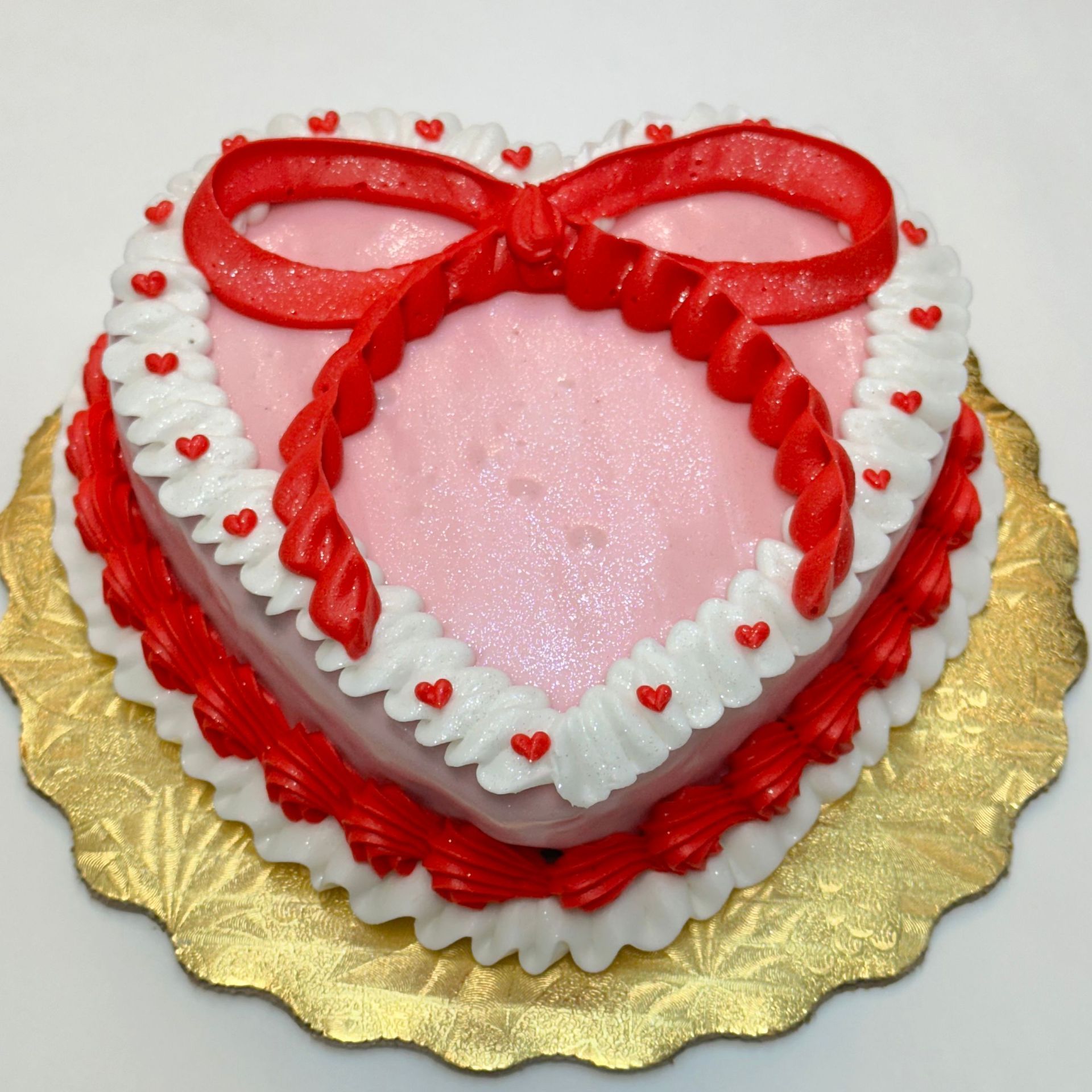 Heart-shaped pink cake with red bow and frosting details, on a gold scalloped plate.