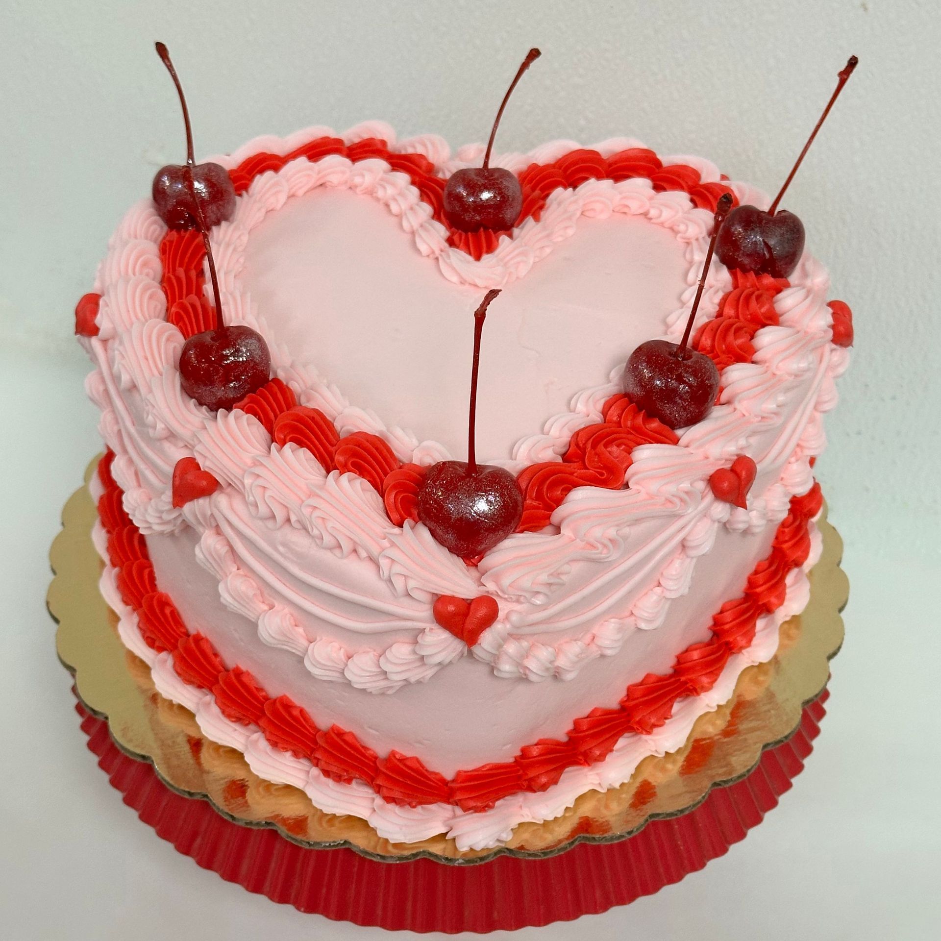 Heart-shaped pink and red frosted cake, decorated with cherries, sitting on a gold and red doily.