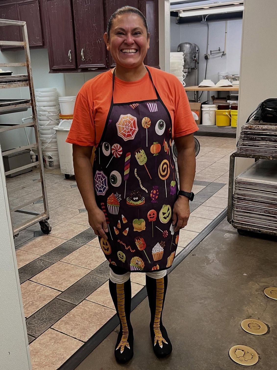 Woman wearing Halloween apron and chicken-foot boots smiles in a bakery setting.