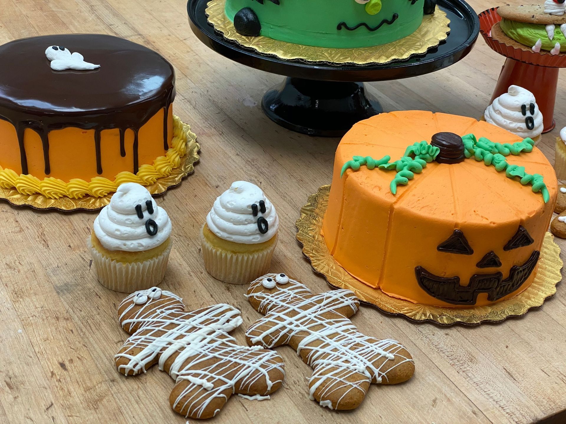 Halloween-themed cakes, cupcakes, and cookies on a wooden table. Pumpkin and monster cakes, ghost cupcakes, gingerbread men.
