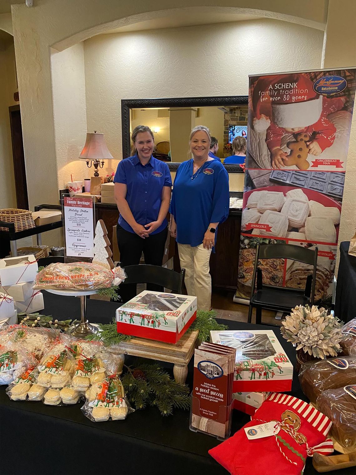 Two women at a table displaying baked goods, Christmas-themed packaging, and a promotional poster.