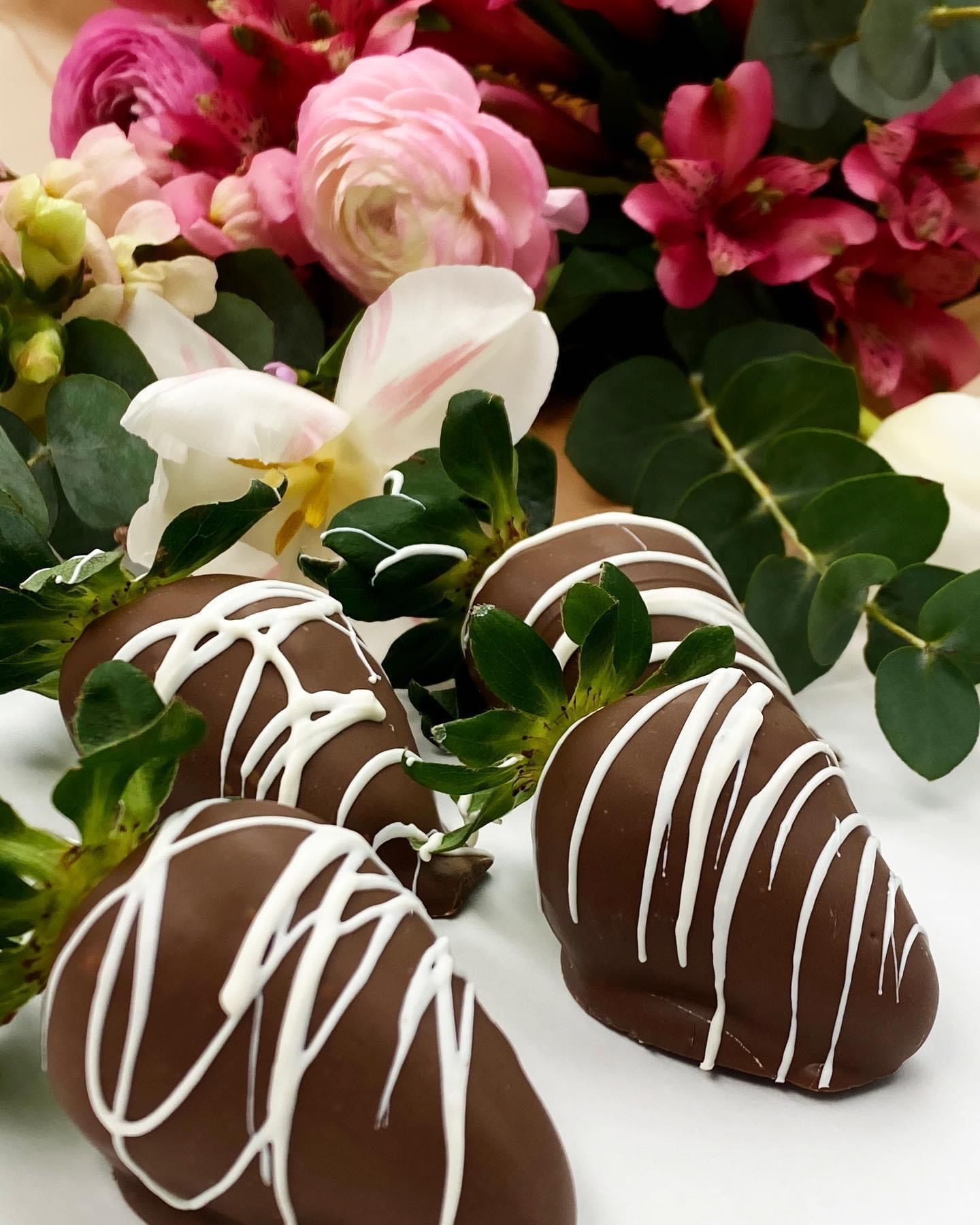 Chocolate-covered strawberries drizzled with white icing, set against a colorful bouquet of flowers.
