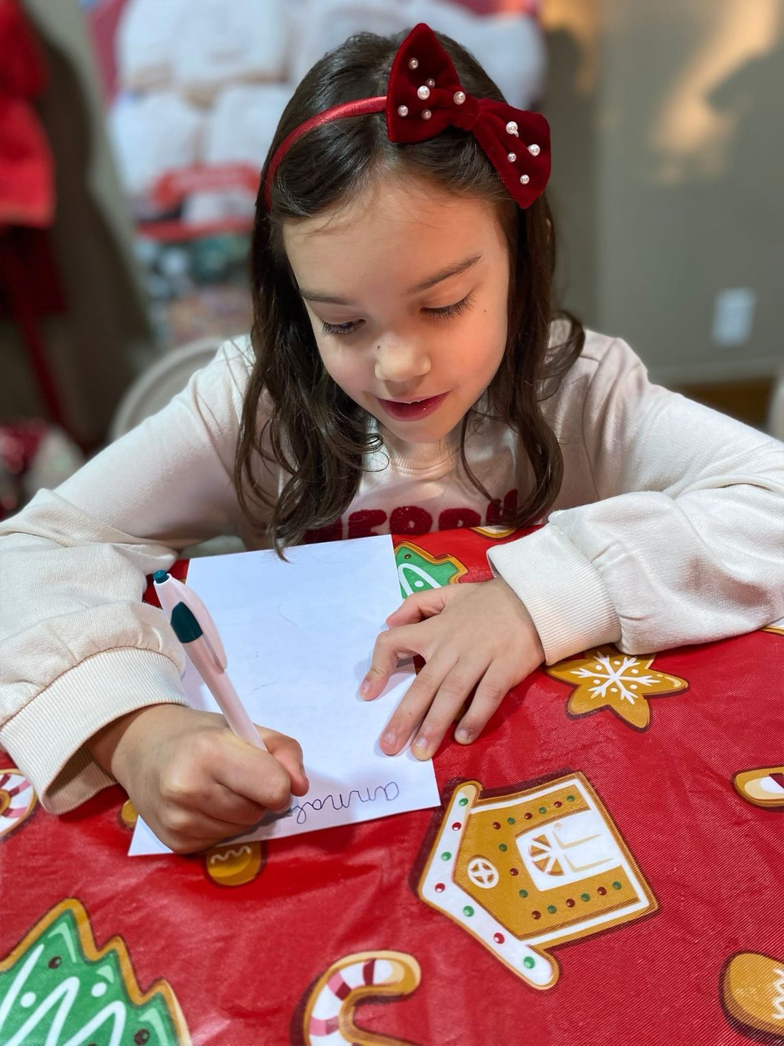 Girl writing on paper at a Christmas-themed table. Red bow headband, pen in hand, festive decor in the background.