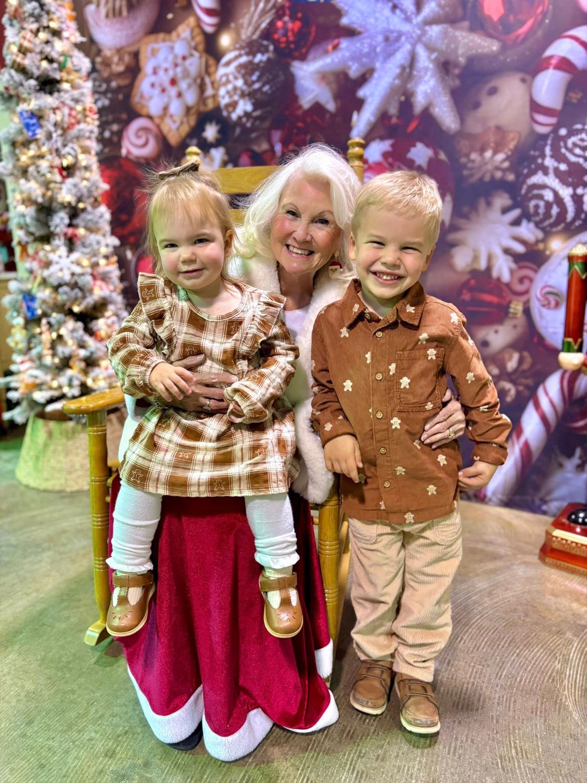 Woman and two children in front of Christmas decorations; smiling, one child on lap, one beside, all in festive outfits.
