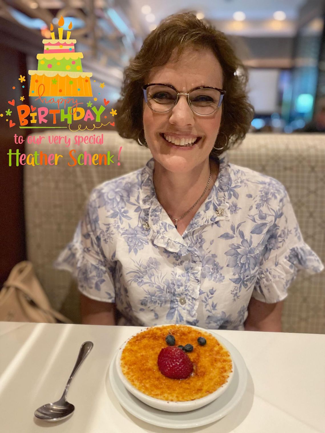 Woman smiles, celebrating birthday with a dessert. She wears glasses and a blue floral shirt, sitting at a table with crème brûlée.