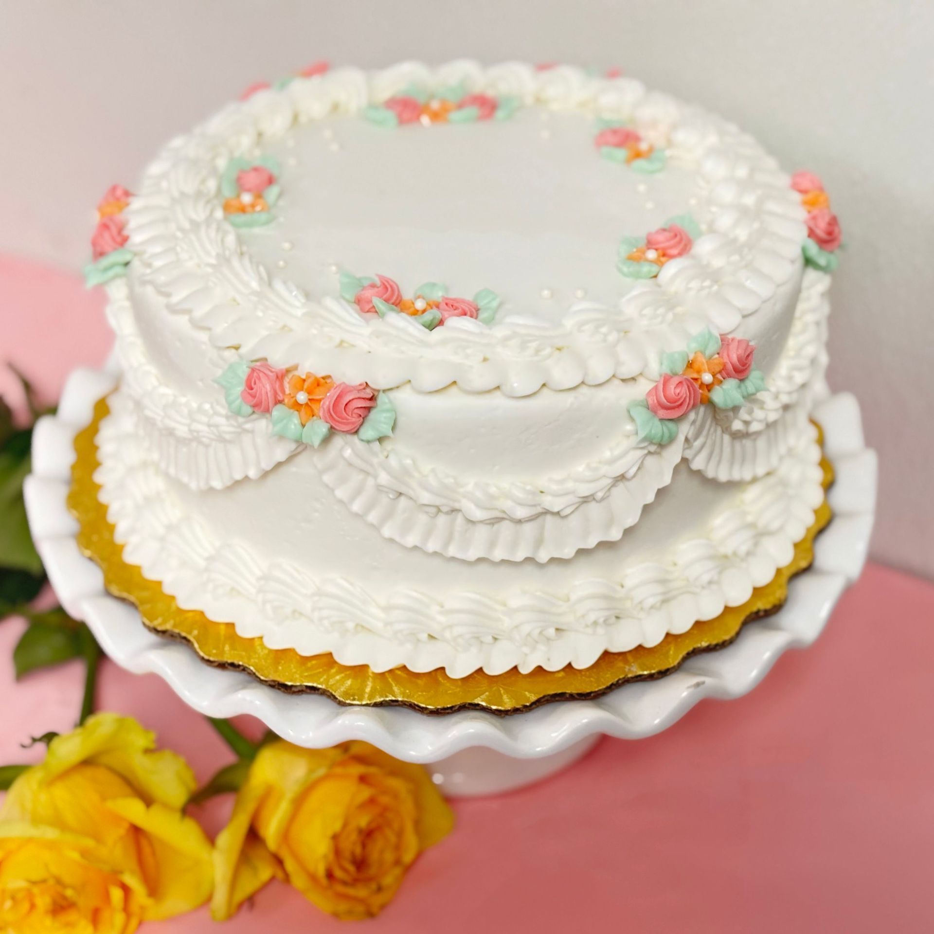 Two-tiered white cake with ruffled frosting, floral accents, on a white pedestal, with yellow roses.