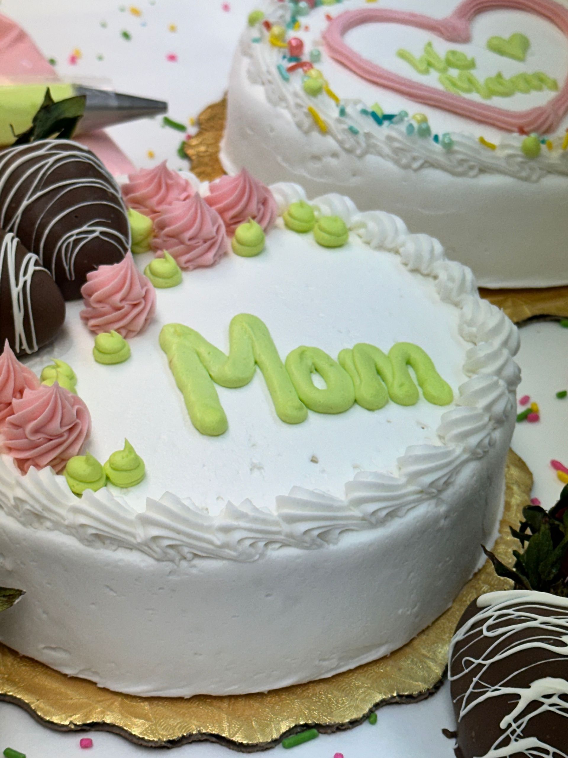 Two round white frosted cakes decorated for Mother's Day, featuring green Mom lettering, pink rosettes, and strawberries.
